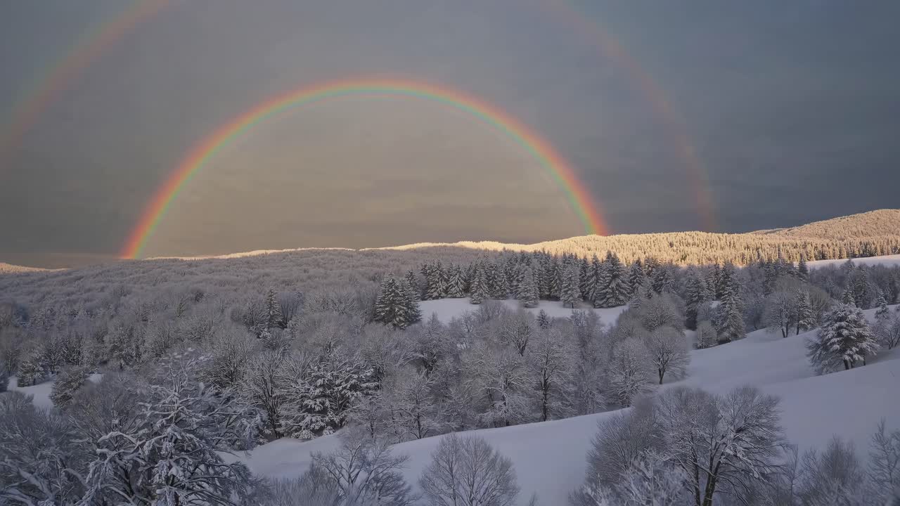 Aerial view of a snowy landscape with a vibrant double rainbow arching over a forest