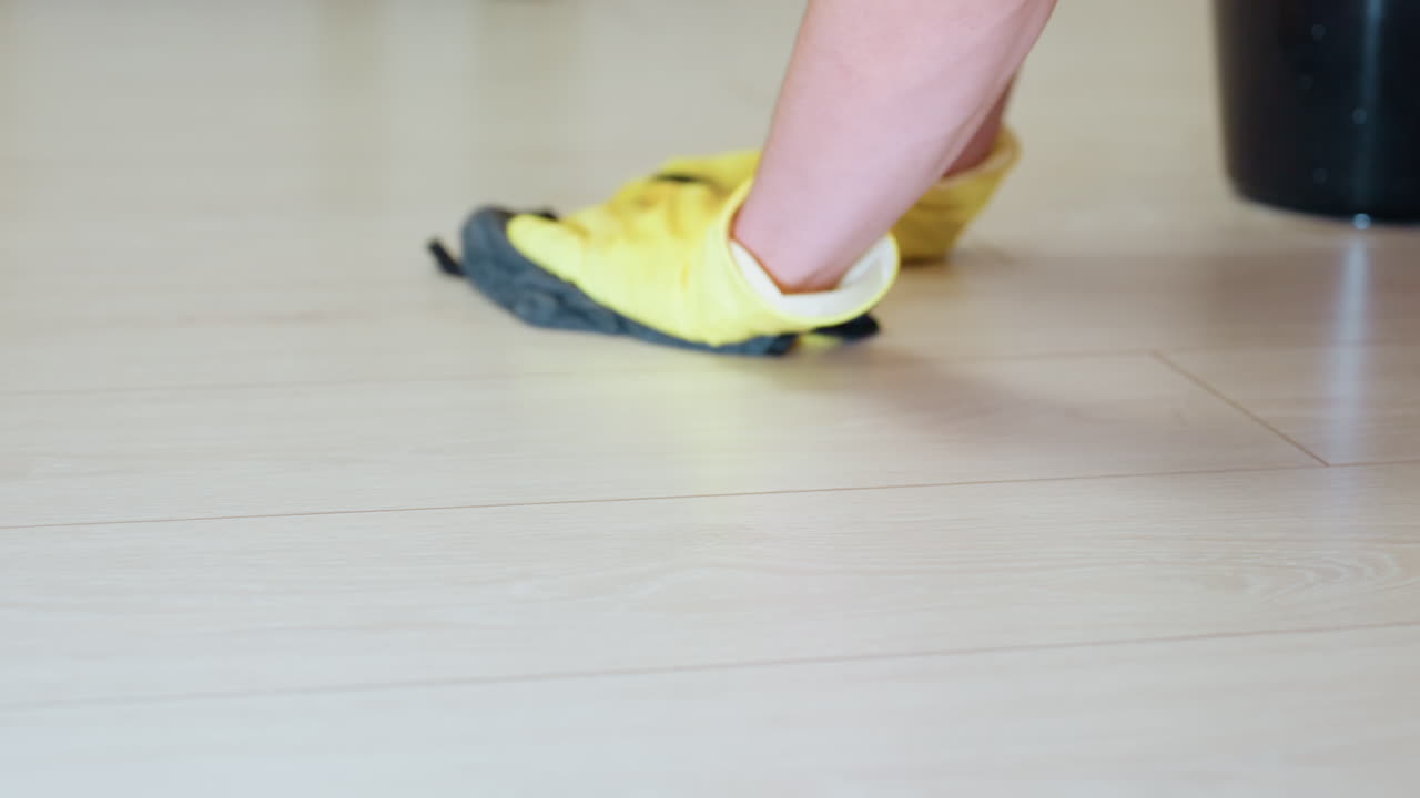 Close up hand view of person wearing yellow gloves scrubbing wooden floor with black cloth, highlighting detailed movement, cleanliness effort, and careful domestic house cleaning work