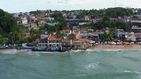 Rotating-aerial-drone-shot-approaching-the-beautiful-pink-Saint-Sebastian-Chapel-in-the-famous-tropical-tourist-beach-town-of-Pipa,-Brazil-in-Rio-Grande-do-Norte-on-a-warm-sunny-summer-evening