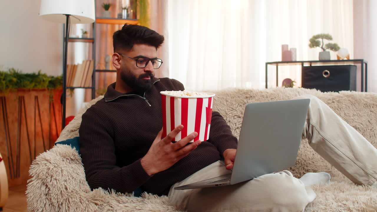 Indian man watching movie on laptop and eating popcorn at home smiling and enjoying leisure time