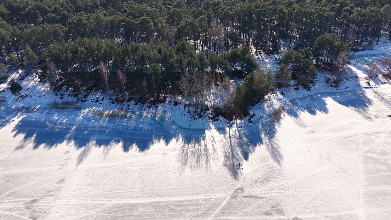 Un'inquadratura aerea mozzafiato cattura un sereno paesaggio invernale con un bordo di foresta coperto di neve che incontra un vasto lago ghiacciato. Il sole proietta lunghe ombre, creando un contrasto drammatico.