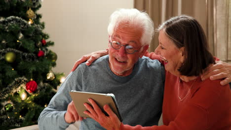 senior couple laughing together at home, using tablet near christmas tree