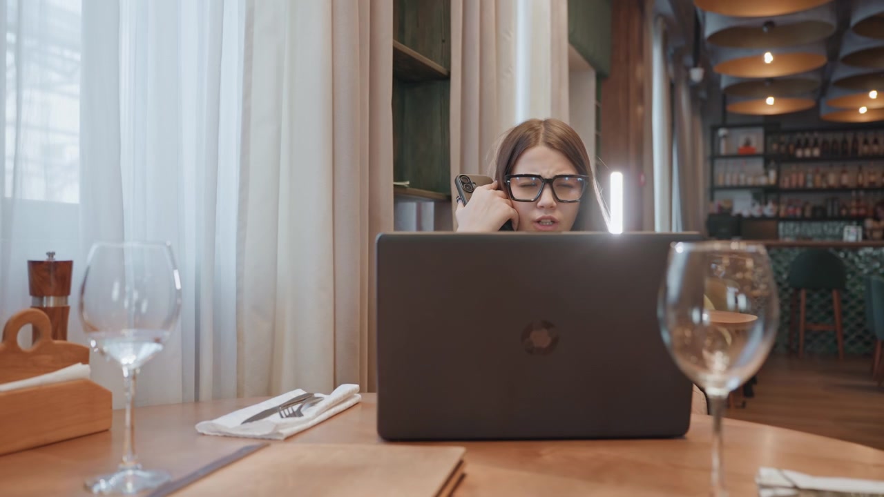 Caucasian woman typing on laptop in restaurant, holding phone to ear while checking messages, tense freelancer negotiating meeting, wine glass in foreground, daylight filtering through curtains