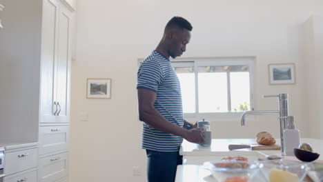 happy african american man pouring into cup and drinking coffee in sunny kitchen, slow motion