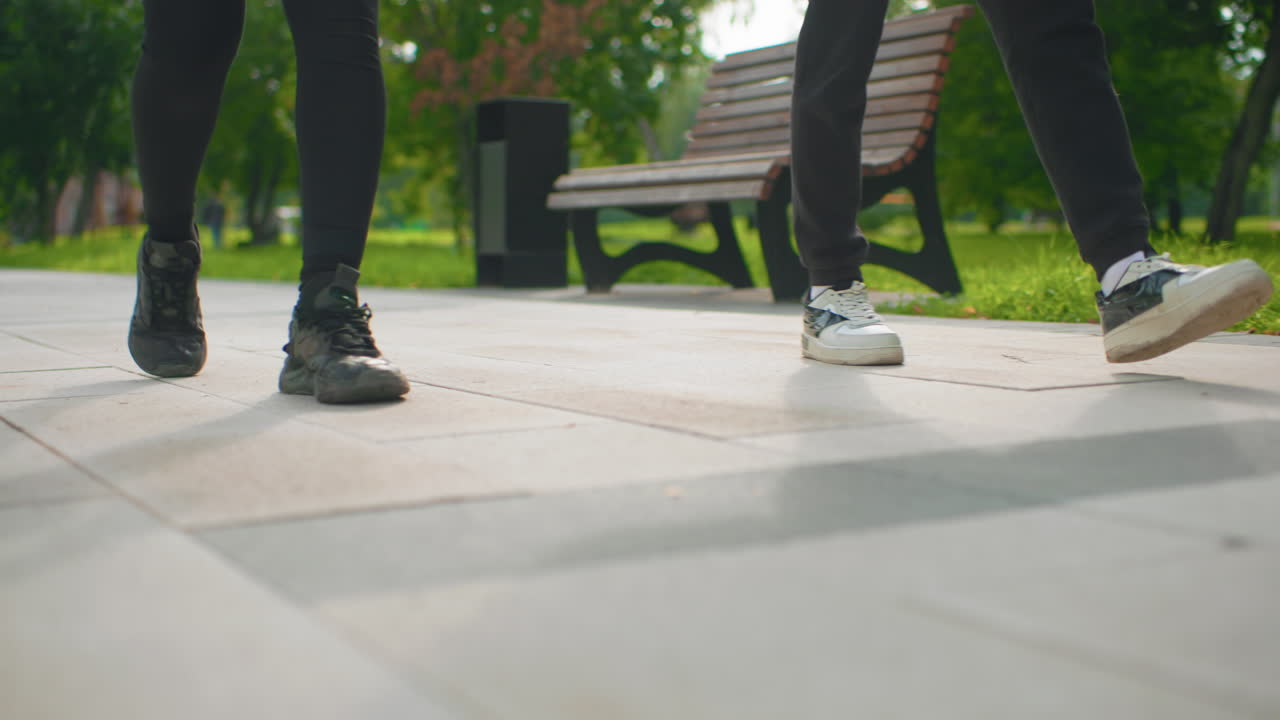closeup sneakers pacing on park sidewalk, two training partners step in sync past benches and trees, morning light, focused cadence and steady pace, athletic energy and toetotile contact captured