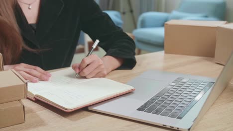 young asian transgender woman business owner with boxes on the table happy online sales job, get an order from customers, use laptop computer and notes on notebook