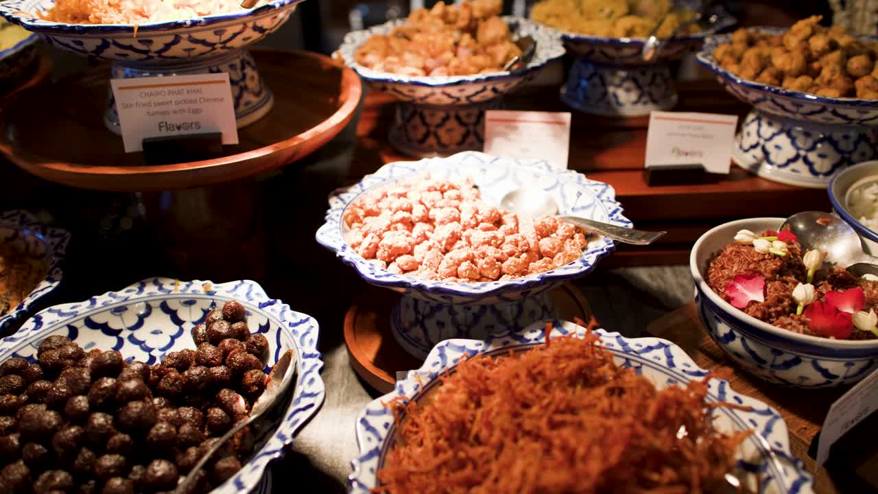 La telecamera effettua una panoramica su un variegato buffet internazionale che presenta cucina tradizionale thailandese e petali di rosa decorativi.