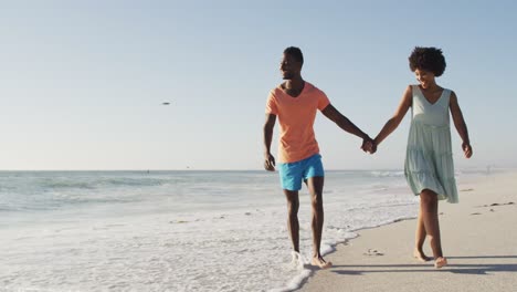 Smiling-african-american-couple-holding-hands-and-walking-on-sunny-beach