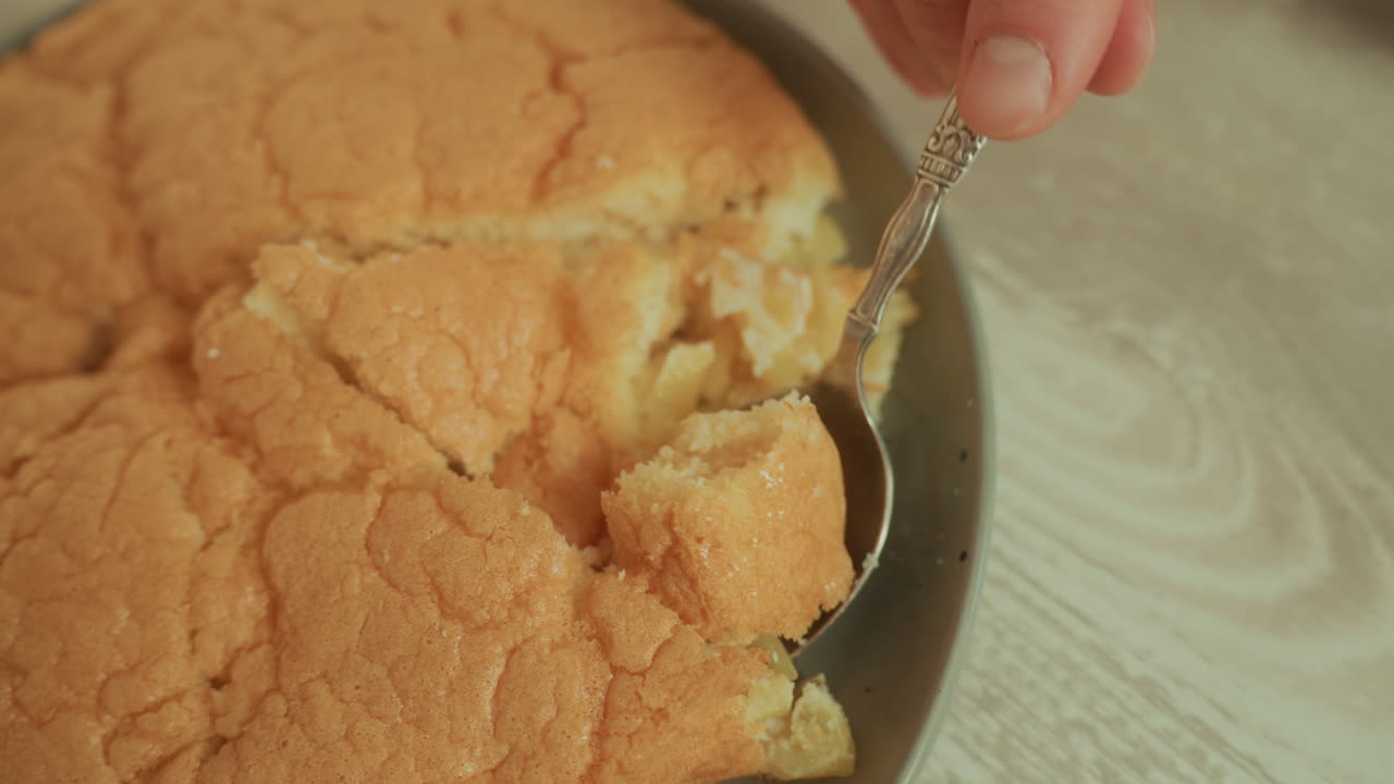 Pan di Spagna dorato prelevato da un piatto con un cucchiaio, mollica soffice e interno umido. Primo piano della superficie testurizzata, dessert fatto in casa su un tavolo di legno, luce naturale calda, atmosfera di cucina rustica e accogliente.