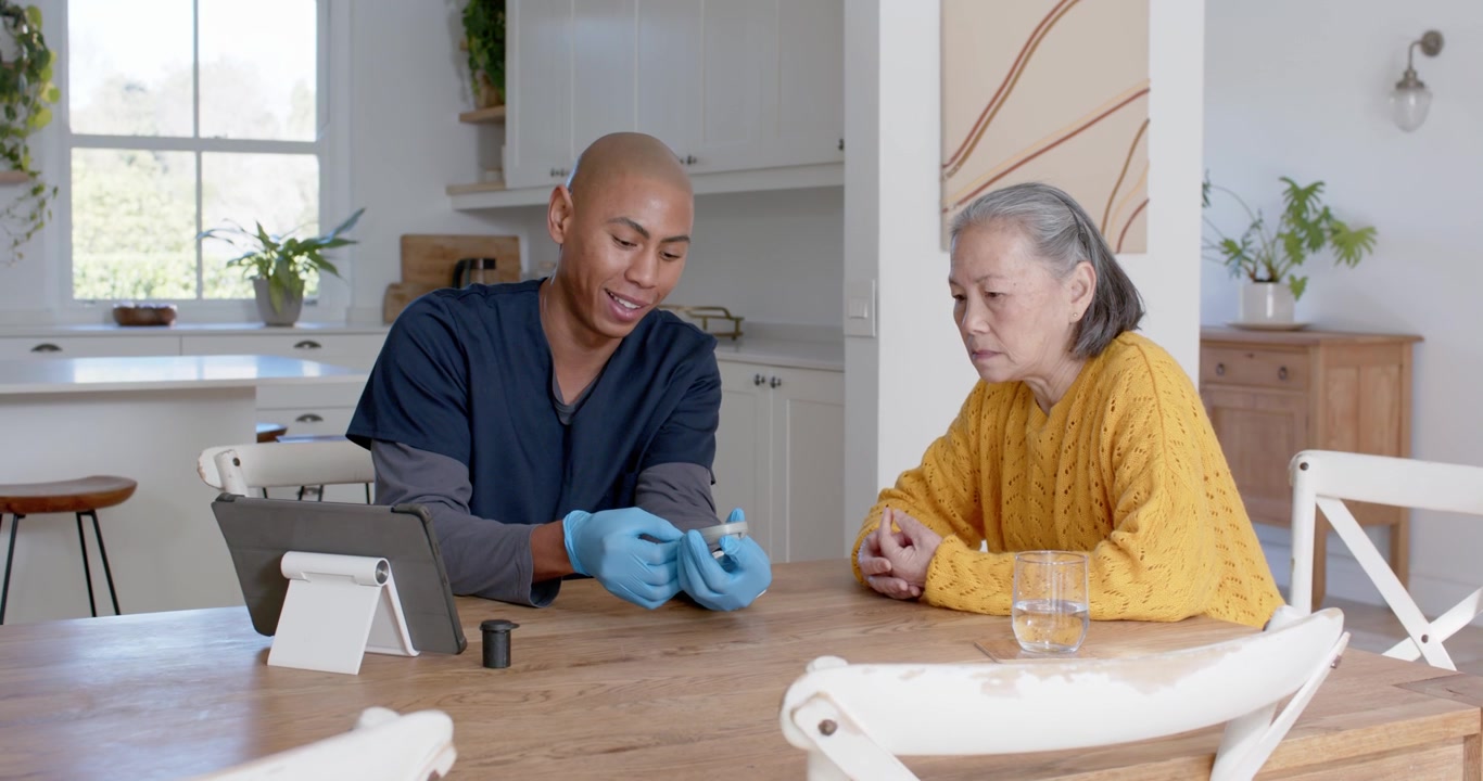 Caregiver in scrubs demonstrating glucose meter at dining table via tablet teaching Senior woman