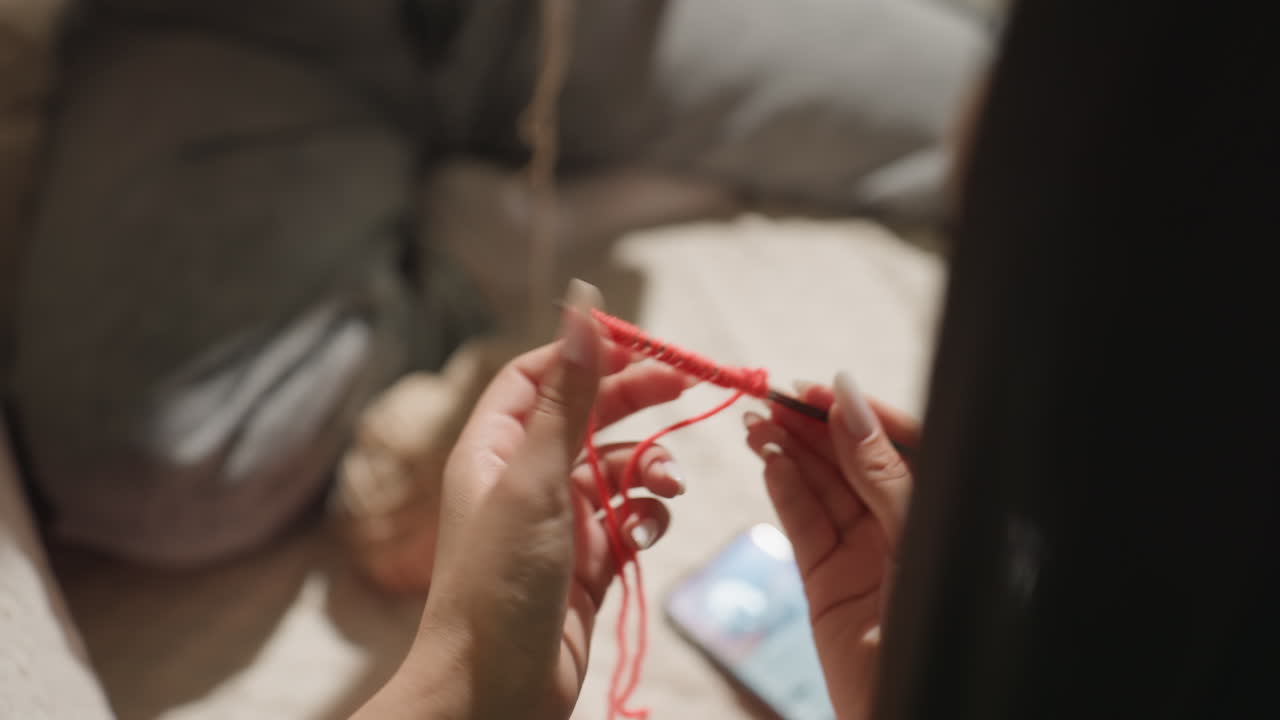 아늑한 거실 안에서 빨간색 털실로 바늘뜨기를 하는 백인 손의 클로즈업, 기본 스티치 연습하는 학생, 고리를 만드는 집중된 손가락, 부드러운 조명, 튜토리얼 분위기, 느리고 신중한 창작