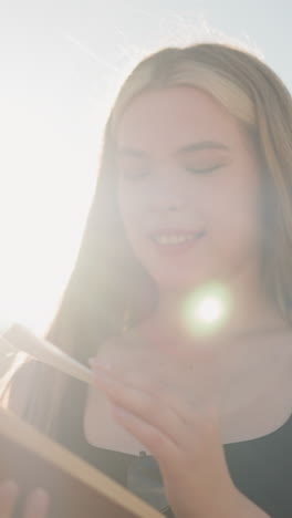 primer plano de una joven vestida de negro hojeando un libro al aire libre bajo un cielo brillante, encontrando papel escondido entre las páginas, mirándolo con curiosidad, sonriendo suavemente mientras la luz del sol ilumina suavemente su rostro