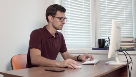 joven empleado de oficina con gafas sentado en la oficina junto a la ventana y escribiendo un informe en la computadora. teléfono inteligente en la mesa. filmado en 4k