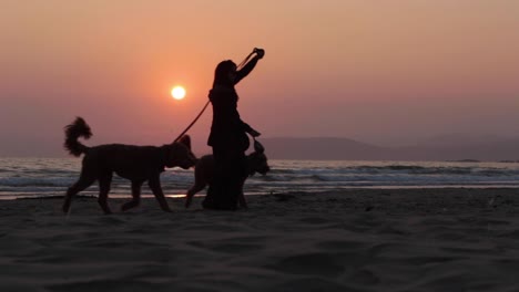 Sonnenuntergang-über-Einer-Inselsilhouette-Und-Ruhigem-Rauschendem-Ozean-An-Einem-Sandstrand,-Während-Eine-Frau-Mit-Zwei-Hunden-An-Der-Leine-Am-Pismo-Beach-In-Kalifornien-Spazieren-Geht