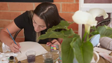 woman hunches over table working intently to watercolor paint a beautiful art piece