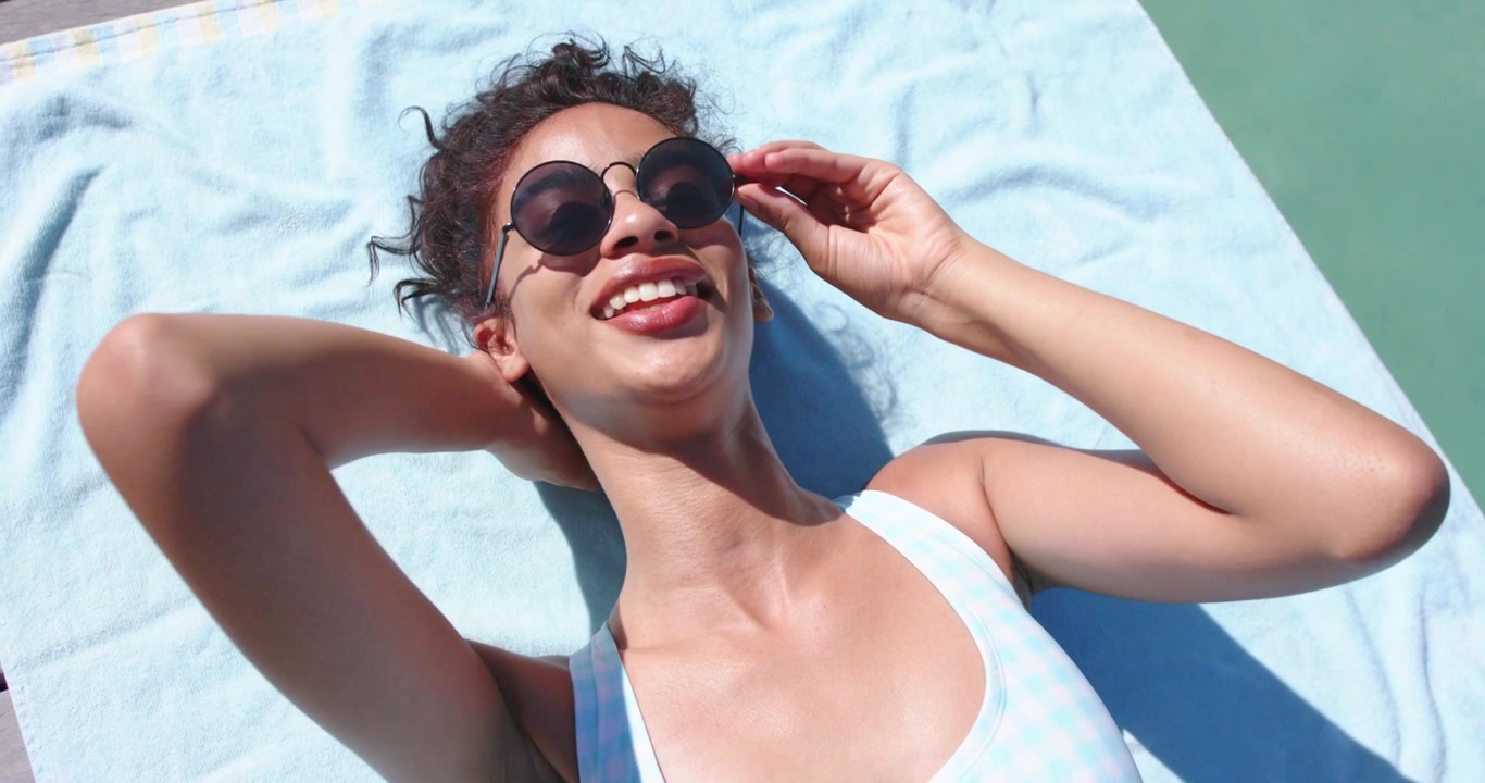 African American adult woman reacting to bright sun, adjusting round sunglasses on light blue towel