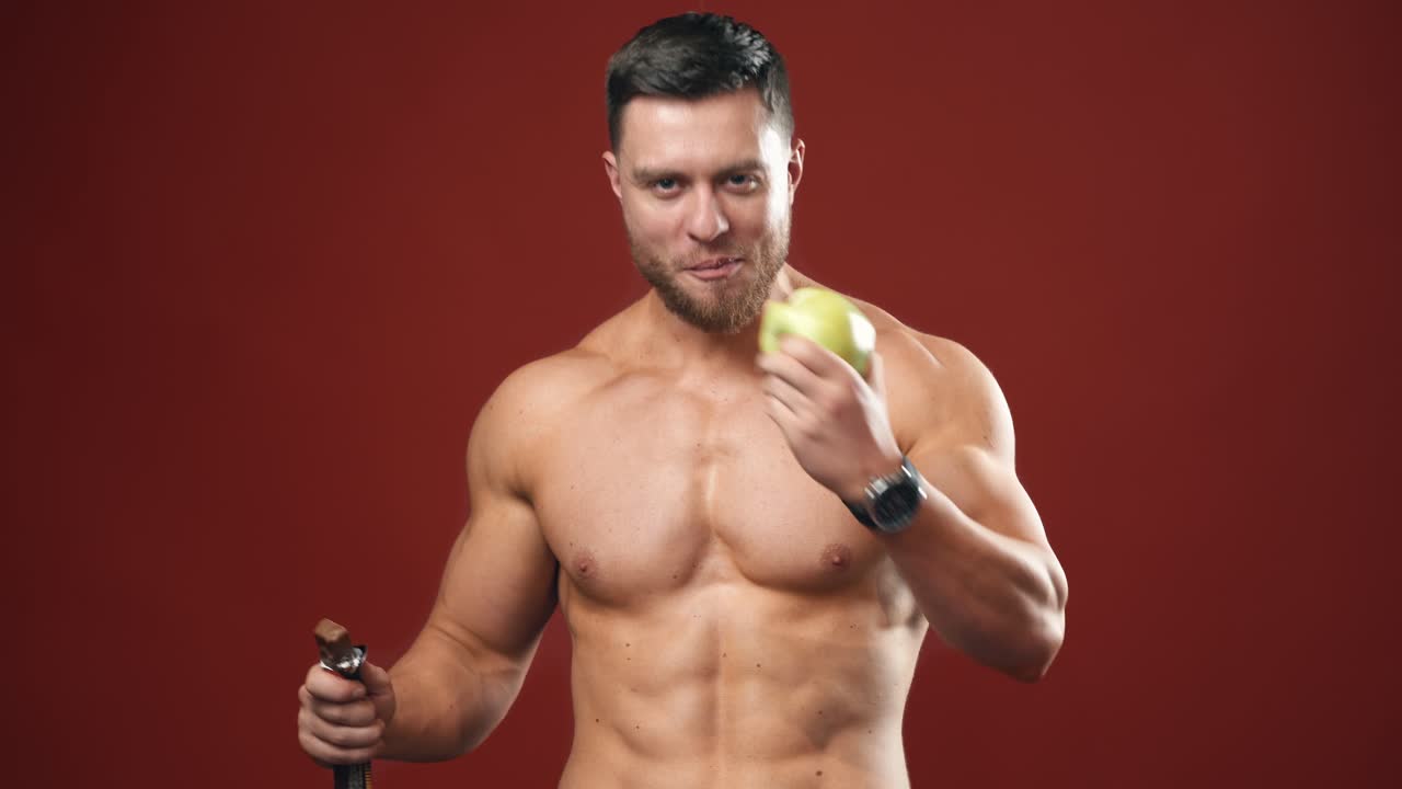 Shirtless bodybuilder eating apple. Portrait of a handsome man with muscular body chooses fresh fruit instead chocolate bar. Smiling strong male eating vitamins in studio.