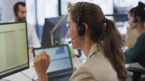 mujer de negocios caucásica sentada en el escritorio hablando usando auriculares de teléfono y computadora en una oficina ocupada