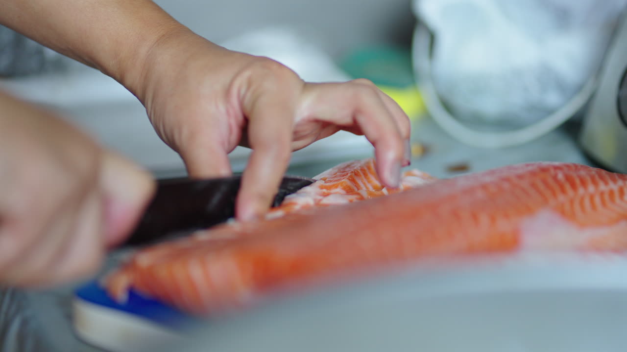 Sushi master expert hands guide yanagiba knife through a fresh salmon fillet in slow motion, carving smooth even sashimi slices for maki rolls on a blue‐rimmed cutting board in a focused kitchen scene