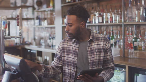 male bar worker standing behind counter checking stock using digital tablet