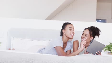 una feliz pareja lesbiana biracial acostada en la cama usando una tableta juntos, en cámara lenta