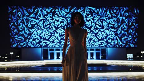 elegant woman in a long dress is confidently walking in front of a modern building illuminated with intricate blue led light patterns at night