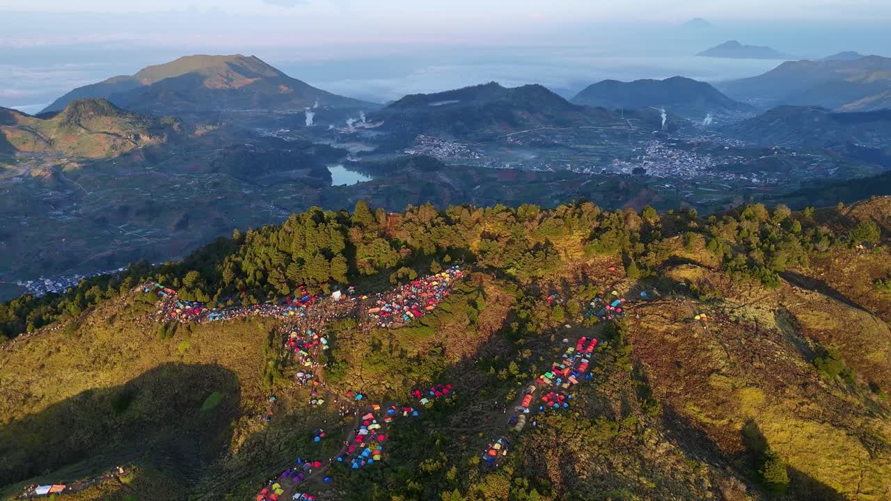 Drone view of densely packed mountain campsite glowing under the morning sun with valleys and distant mountain ranges on background. Sunrise camp point of Mount Prau, Indonesia