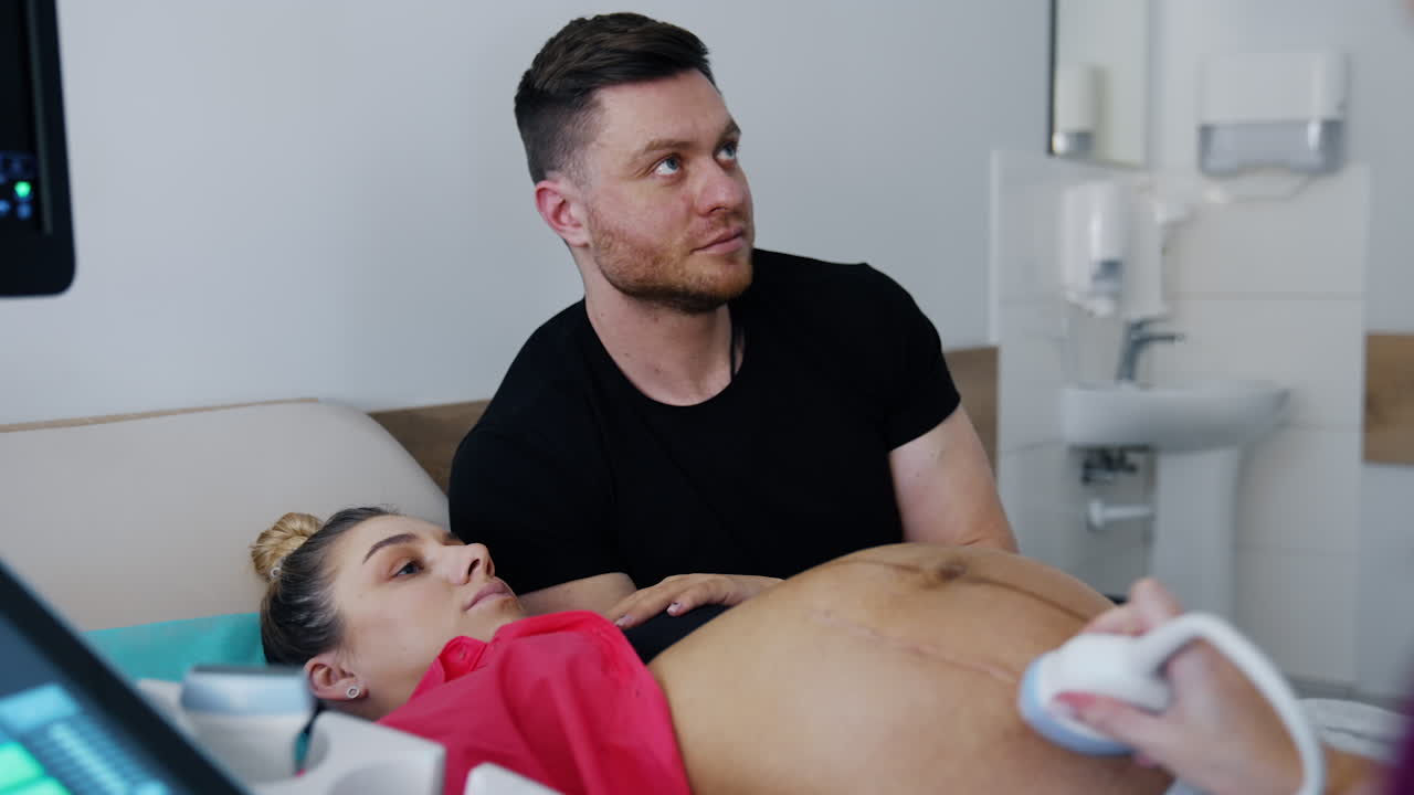 Focused woman expecting a baby looks attentively on the screen of ultrasound check up. Man sits beside his pregnant wife.