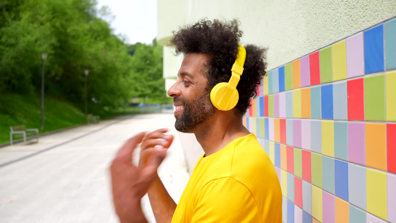Man with yellow headphones enjoys music outdoors next to a colorful wall