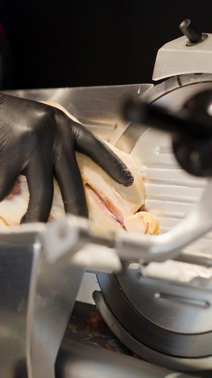 Close-up of gloved hand operating professional meat slicer in commercial kitchen with bright lighting.