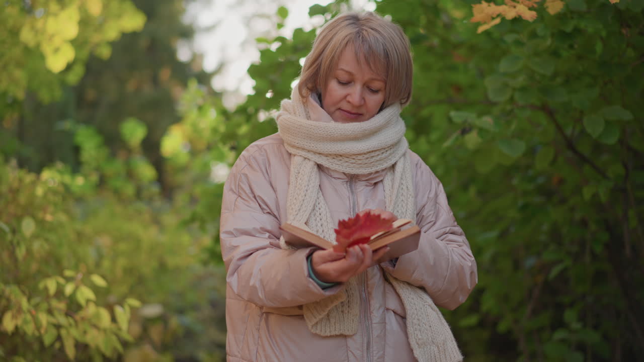 book lover flipping page with autumn leaf in hand while walking through forest, golden foliage framing path as warm sunlight filters through trees, cozy jacket and scarf