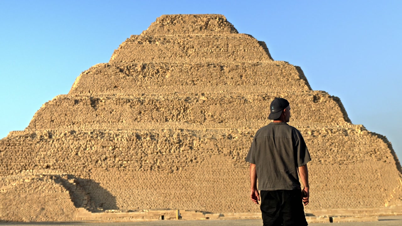 Man walking across the desert plateau of Saqqara as the sun sets, casting a warm, golden glow over the ancient landscape. In the background stands the iconic Step Pyramid of Djoser.
