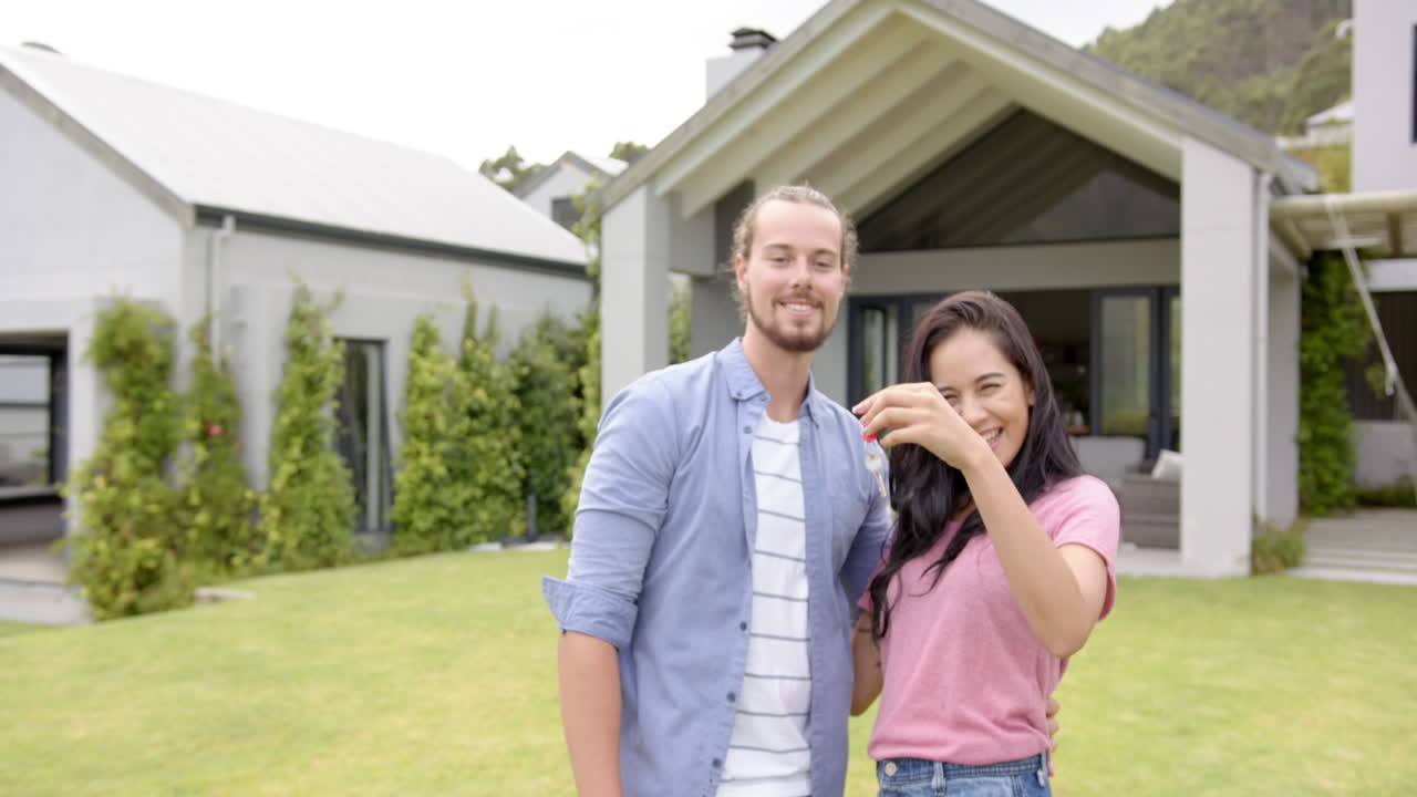 una joven pareja diversa está sonriendo al aire libre en el patio trasero de su casa, sosteniendo llaves