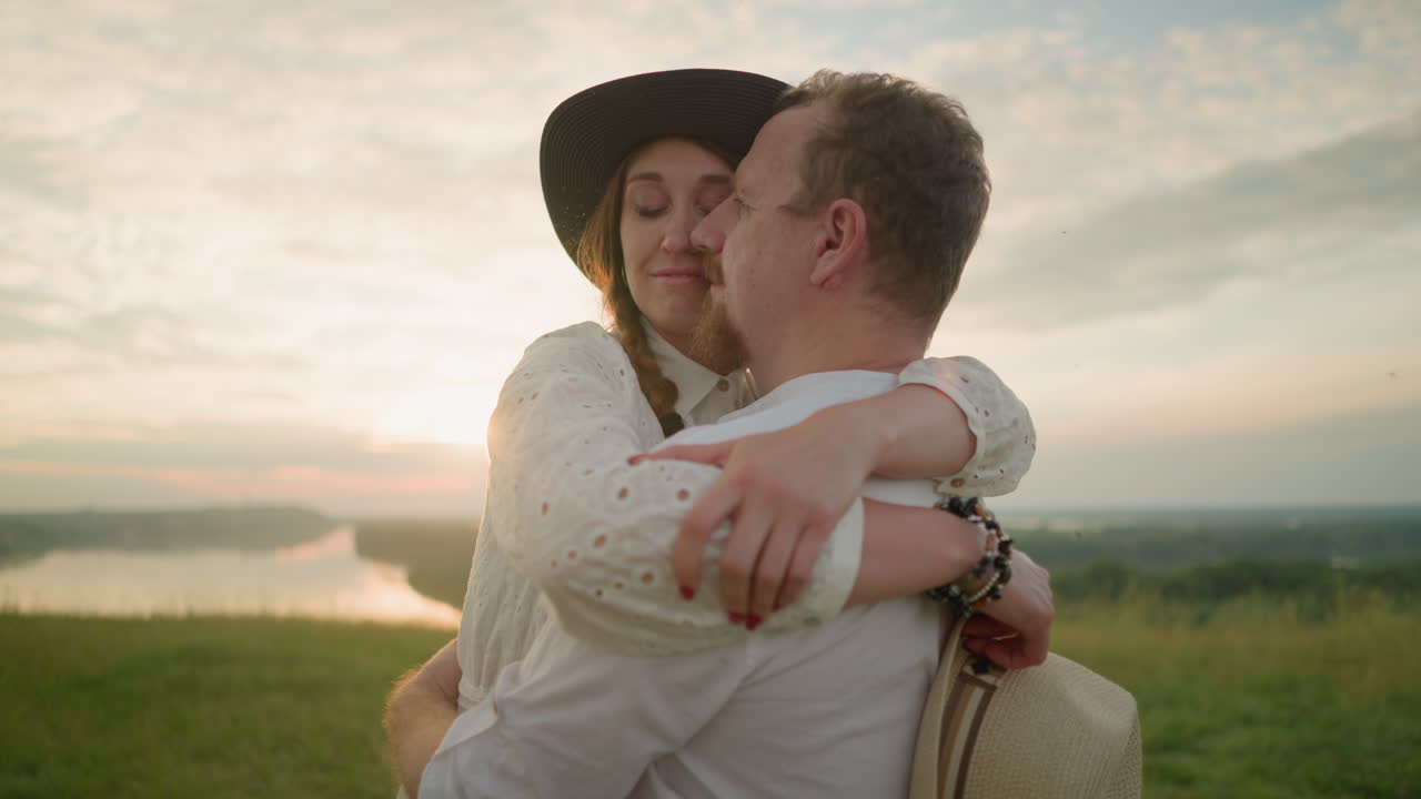 A husband and wife share a tender embrace in a grassy field at sunset, smiling as their faces touch. Both dressed in white outfits, the woman wears a hat and holds another in her hand