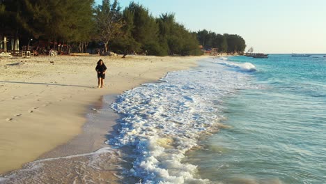 girl walking alongside white waves of blue sea on white sand of exotic beach of tropical island in bali