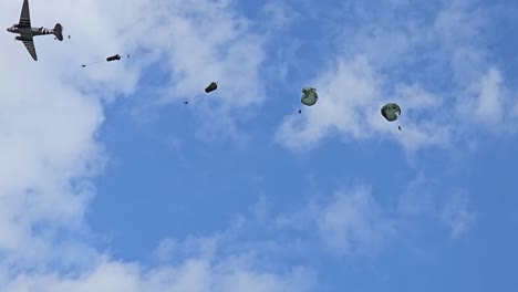 a military aircraft drops parachutes and supplies mid-flight against a blue sky with scattered clouds during an air demonstration.