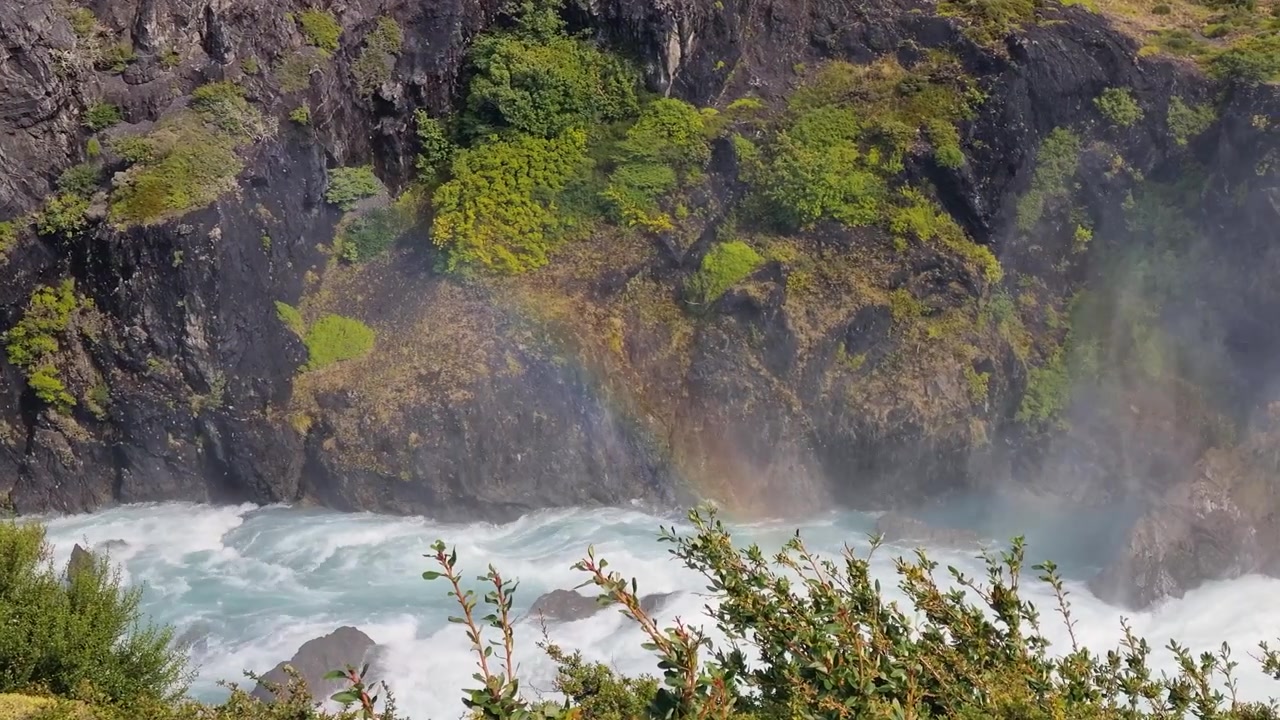 Air Terjun Salto Grande di Taman Nasional Torres del Paine, Chili, dengan air pirus yang deras mengalir melalui lanskap Patagonia saat pelangi cerah melengkung di atas air terjun