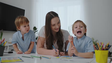 A-young-mother-with-two-children-sitting-at-a-white-table-draws-colored-pencils-on-paper-in-slow-motion