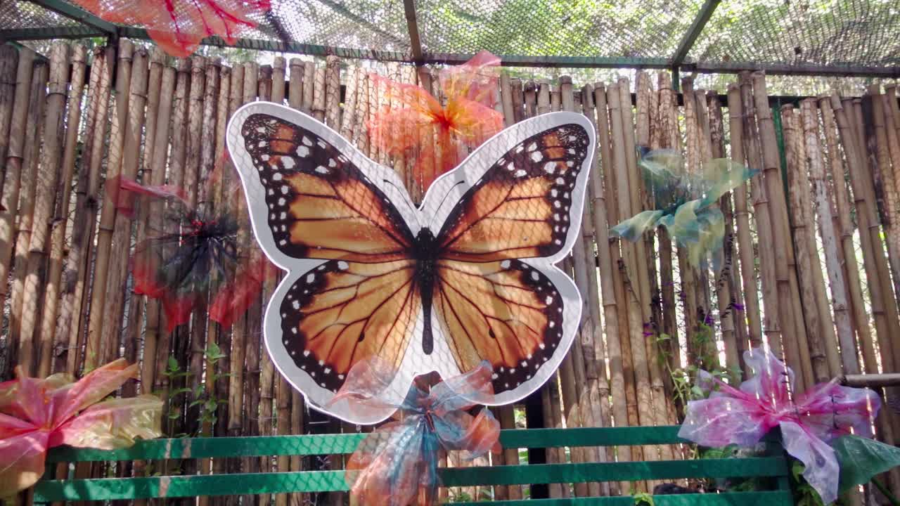 Grande farfalla decorativa montata su una recinzione di bambù con nastri colorati e luce soffusa, scena di decorazione di giardino esterno che crea un'atmosfera artistica vibrante con tema naturale