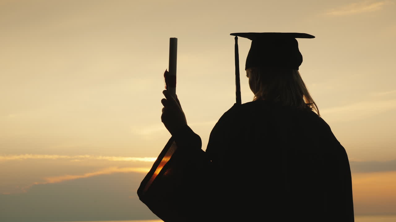 un soltero con un diploma en la mano y una gorra de graduado mira el amanecer sobre el mar