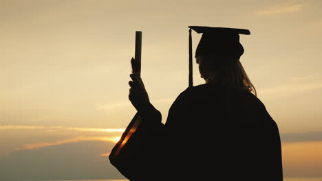 a bachelor with a diploma in hand and a cap of a graduate looks at the sunrise over the sea