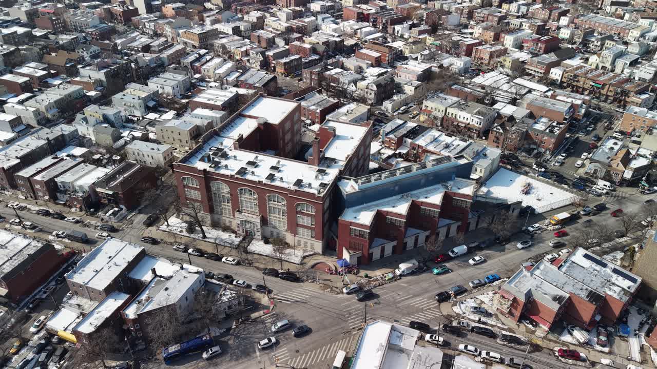 Riprese aeree che si allontanano da un grande edificio scolastico in mattoni a un incrocio, rivelando un vasto quartiere urbano con tetti imbiancati dalla neve e strade cittadine con traffico in movimento