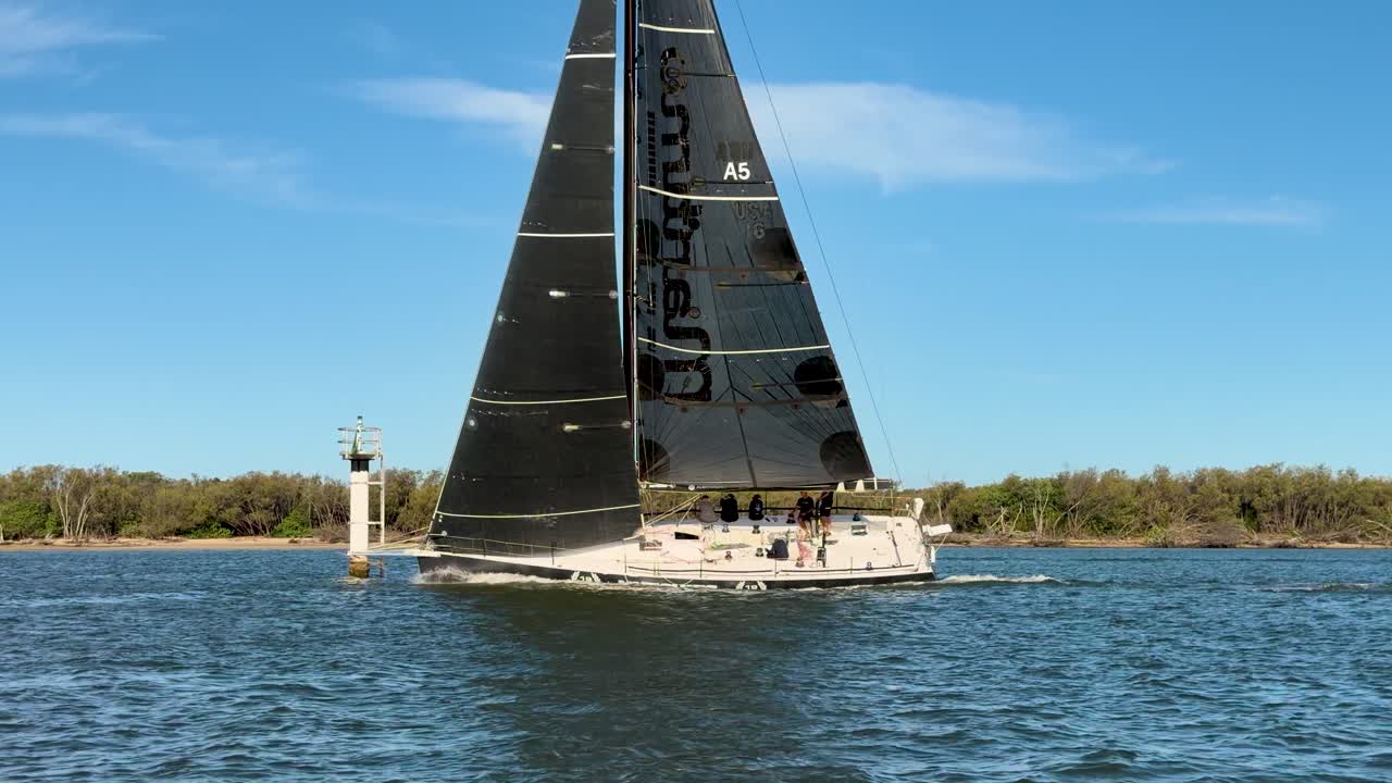 Modern sailboat with black sails glides past navigation marker under clear blue sky, steady camera