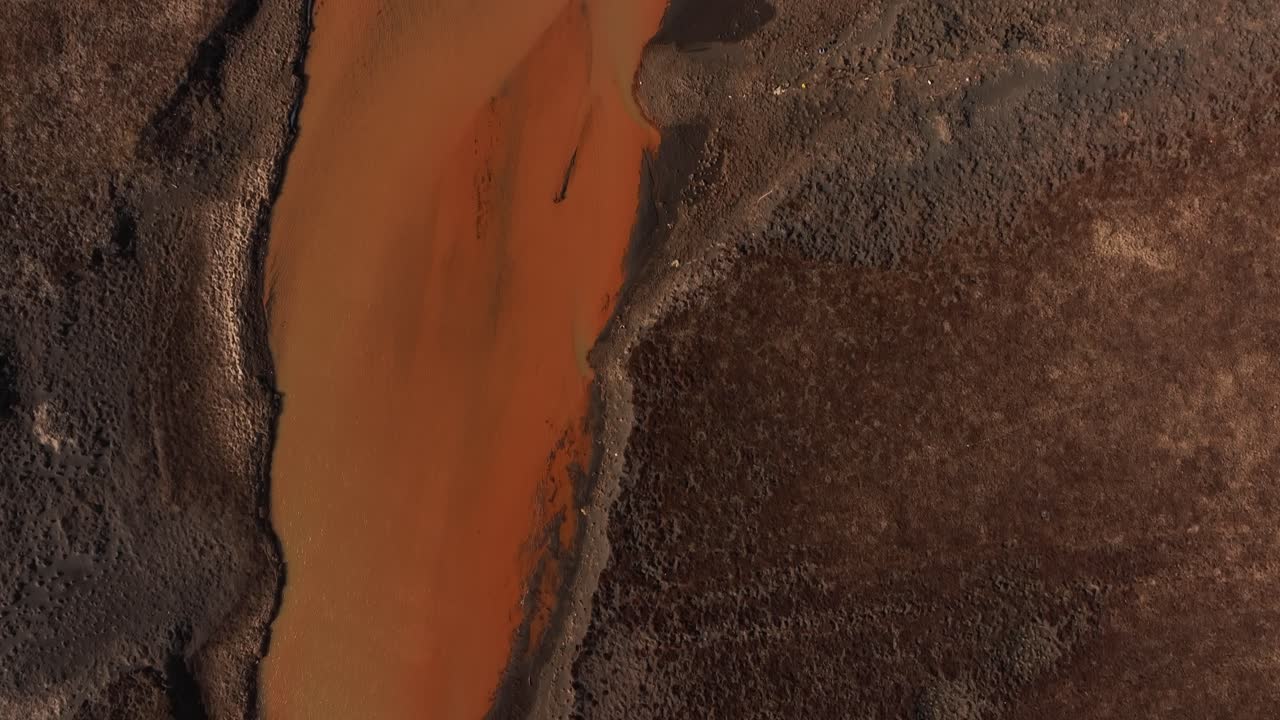 Aerial top-down view of the Yellow River merging with the Þjórsá River in Iceland, creating a striking contrast between the earthy brown terrain and the vibrant orange water.