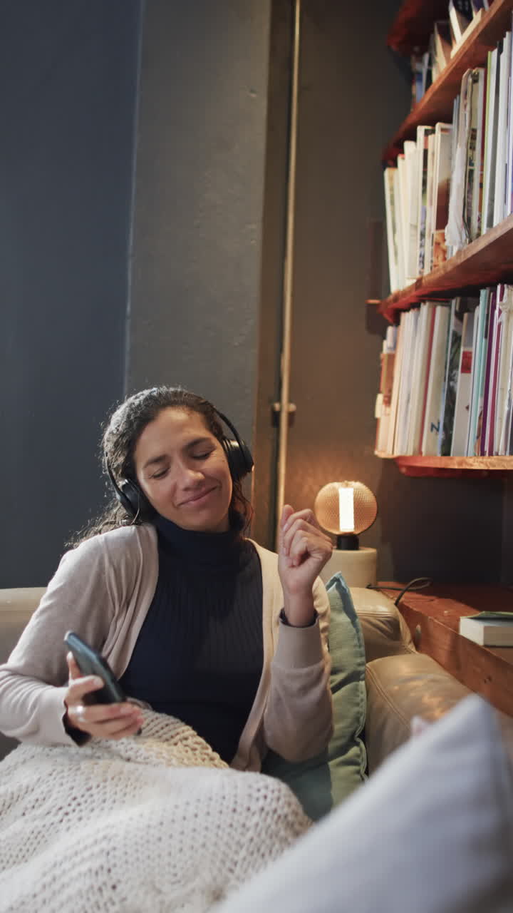 Vertical video of biracial woman listening music using smartphone in living room, slow motion