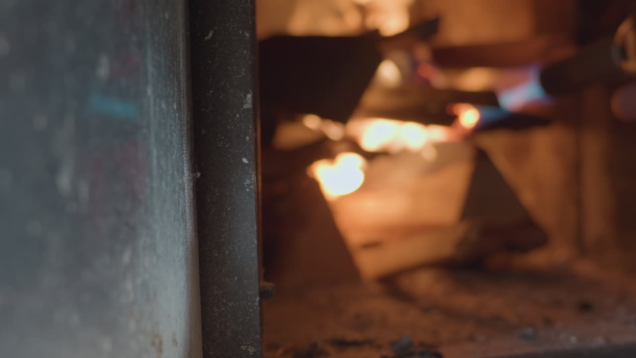 Side view of person using blue flame torch to ignite stacked firewood inside dark indoor fireplace, flames begin to grow and flicker warmly, creating cozy glowing effect on wood surface
