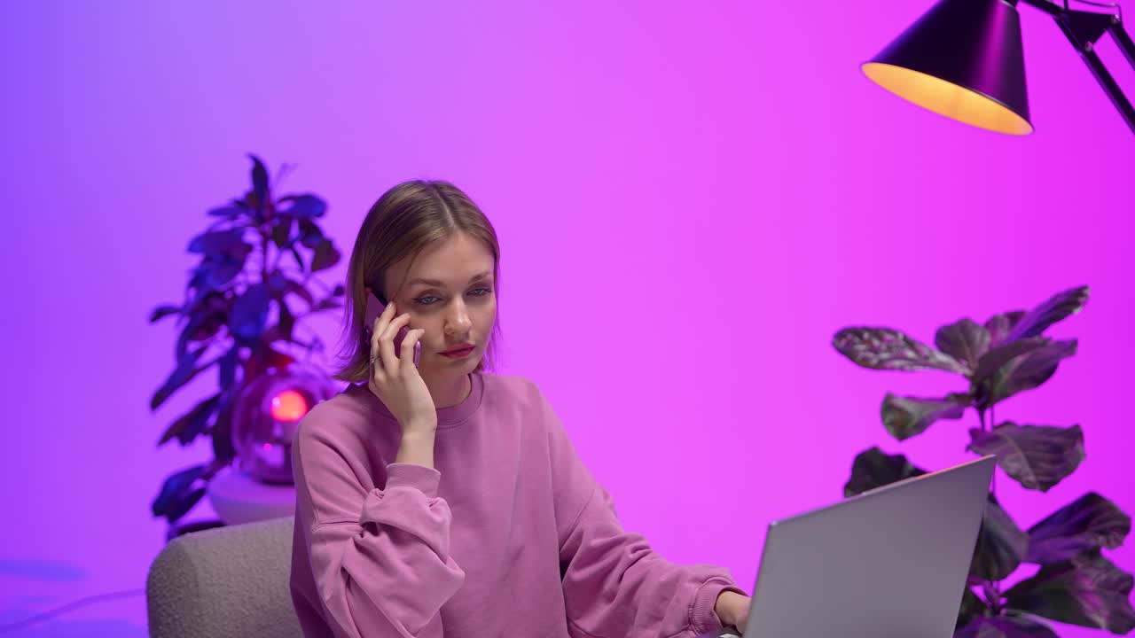 A woman in a pink sweatshirt holds a phone to her ear while working on a laptop. Her intense gaze and the vivid purple background create a bold and emotional scene of modern digital communication