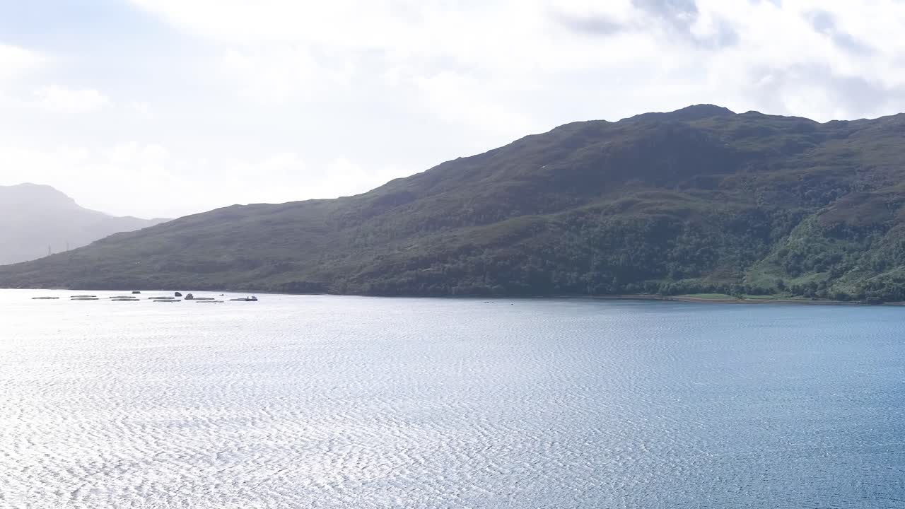 Drone pans across sunlit Scottish Highlands, blue water, forested hills, and distant islands