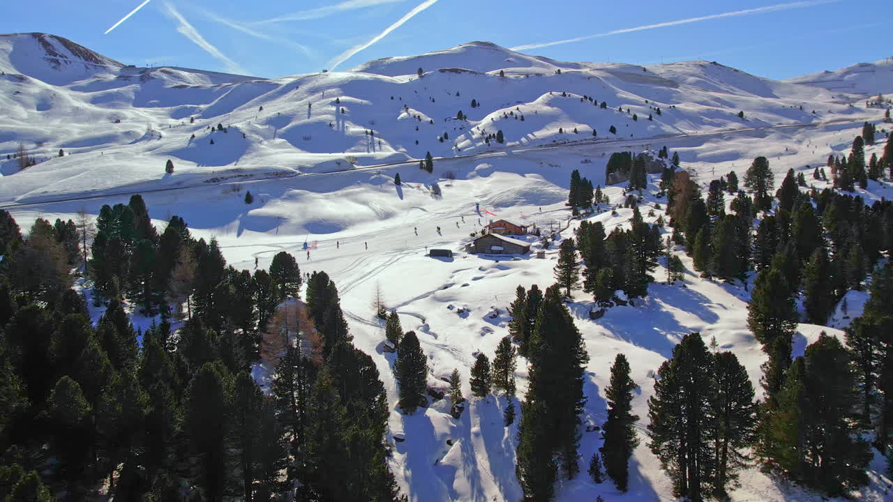 una cautivadora vista aérea de las dolomitas, mostrando a los esquiadores en las laderas nevadas rodeadas de densos bosques de pinos bajo un cielo azul claro