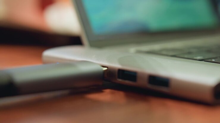 Primo piano di una mano che inserisce una chiavetta USB nella porta di un laptop su una scrivania di legno, luce calda della sera, freelancer concentrato che esegue il backup di file importanti su una memoria esterna, movimento lento e deliberato, profondità di campo ridotta.